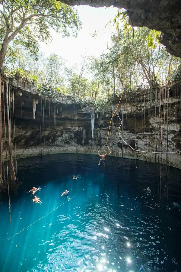 Personne sautant depuis une corde dans l'eau bleu turquoise d'un cénote naturel bordé de lianes.