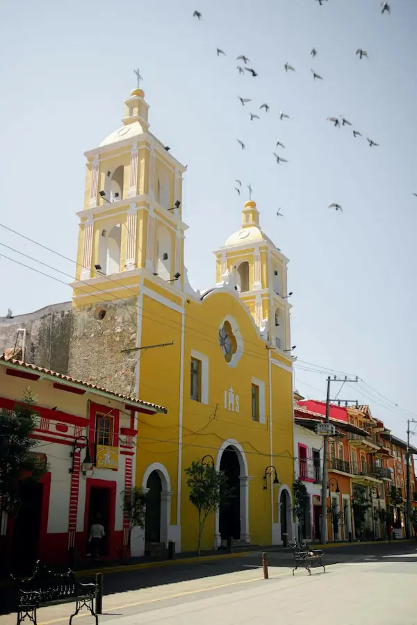 Église coloniale jaune sous un ciel clair avec des oiseaux volant au-dessus d'une rue mexicaine.