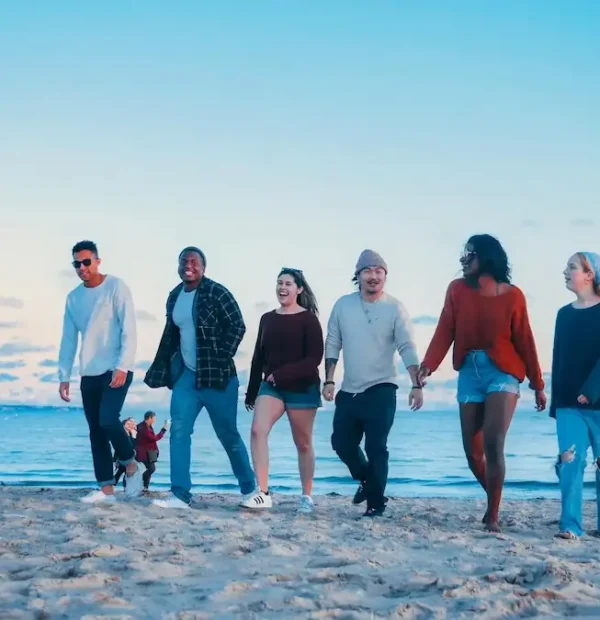 Groupe de jeunes voyageurs marchant et riant ensemble sur une plage de sable en bord de mer.