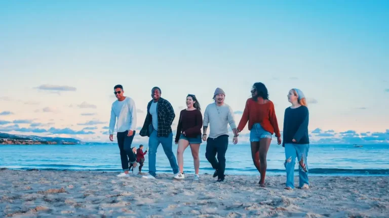 Groupe de jeunes voyageurs marchant et riant ensemble sur une plage de sable en bord de mer.