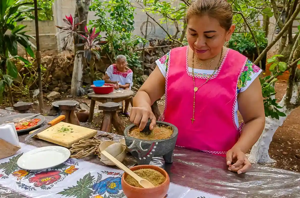 Femme maya écrasant des condiments dans un molcajete en pierre lors d'un repas partagé en extérieur.