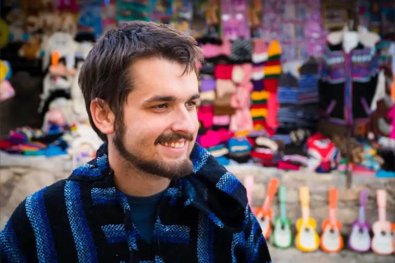 Jeune homme souriant portant un poncho bleu artisanal devant un étal de marché mexicain coloré.