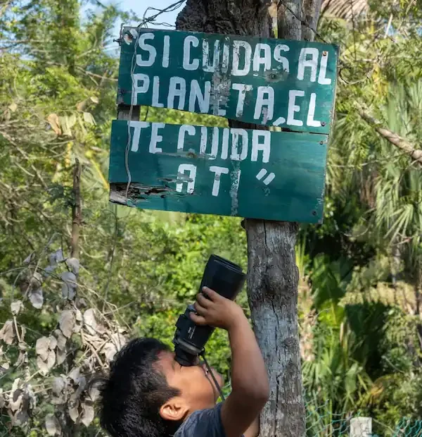 Jeune garçon mexicain regardant à travers des jumelles sous un panneau engagé "Si cuidas al planeta, el te cuida a ti".