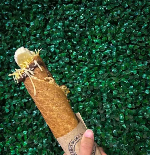 Main tenant une marquesita croustillante garnie de fromage râpé et de banane devant un fond vert.