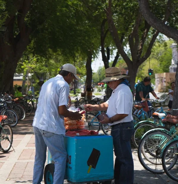 Vendeur mexicain servant un client à son chariot ambulant bleu sous l'ombre des arbres.