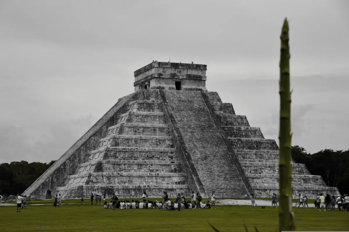 Pyramide de Chichén Itzá au Mexique sous un ciel gris avec un groupe de visiteurs assis sur la pelouse au premier plan.